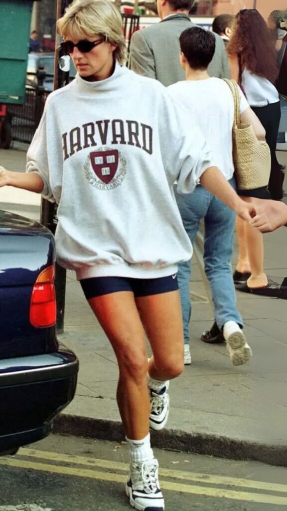 The outfit features an oversized gray sweatshirt with a Harvard logo in maroon, made of soft cotton fabric, and is paired with form-fitting black athletic shorts that end mid-thigh. The look is completed with classic white high-top sneakers and ribbed white crew socks that are slightly scrunched at the ankle, creating a relaxed yet sporty vibe that remains stylish today. This combination captures the carefree spirit of the '90s while still being functional and chic for modern wear.