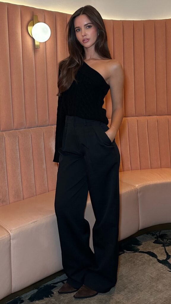 The outfit features a sophisticated one-shoulder, ribbed black sweater, paired with high-waisted, wide-leg black trousers, exuding elegance and style. The textured fabric of the top contrasts with the smooth, tailored look of the pants, creating a chic and polished appearance suitable for special occasions like birthday parties or date nights. Completing the look, the ensemble is styled with pointed-toe black heels, enhancing the overall refinement of this classy all-black winter outfit.