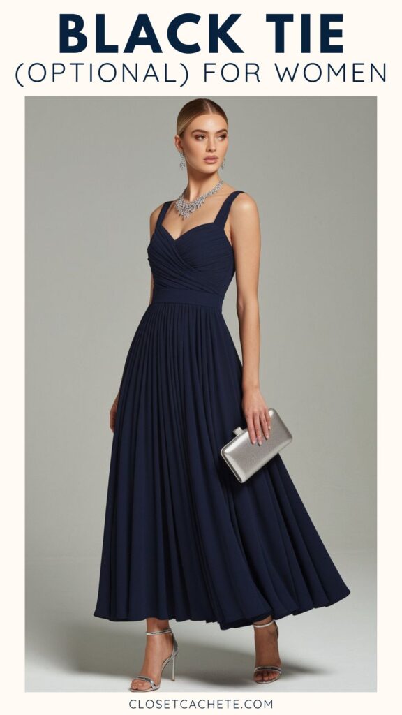 A woman stands elegantly in a floor-length navy blue dress with a fitted bodice and pleated skirt, styled for a black tie optional event. She accessorizes with a sparkling necklace and holds a silver clutch, complemented by strappy silver high-heeled sandals. The neutral background enhances the dress's rich color and the overall sophisticated look.