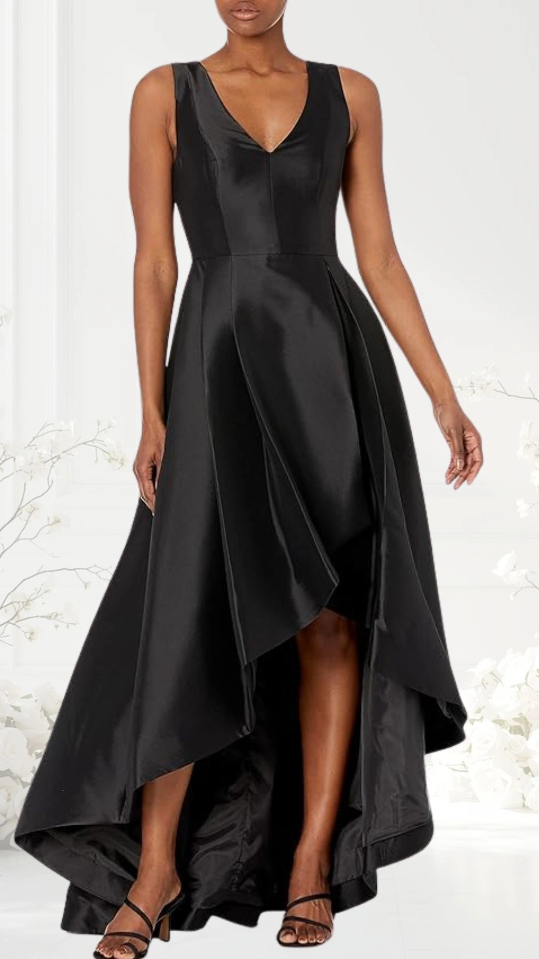 The dress features a sleek black satin fabric with a deep V-neckline and sleeveless design. It has an asymmetric hemline, with a shorter front and flowing back that creates elegant movement, complemented by strappy black heels. The silhouette fits snugly at the waist, enhancing a flattering shape while maintaining a sophisticated look suitable for wedding guests.