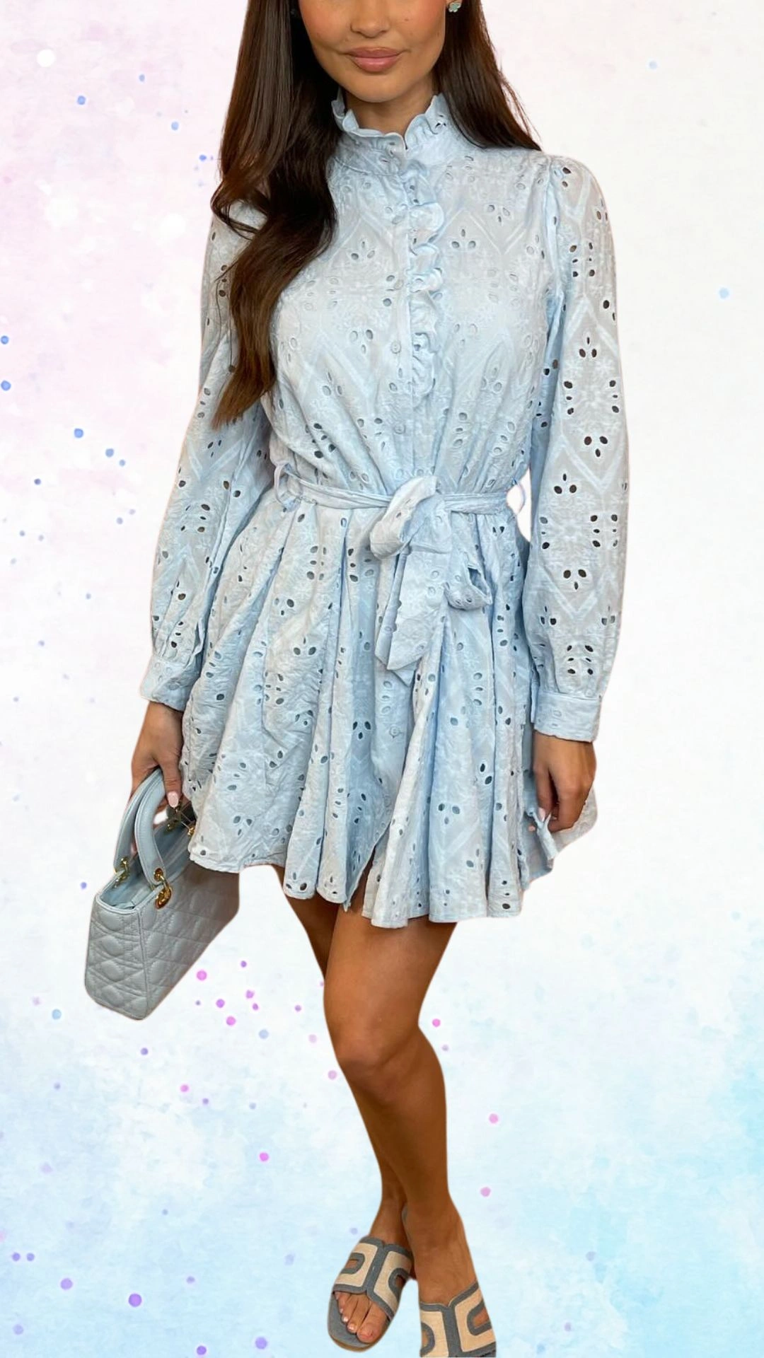 The outfit features a light blue cotton dress with a broderie anglaise texture, characterized by delicate cut-outs and embroidered patterns. It includes long sleeves, a high neckline with button details, and a cinched waist tied with a matching belt, flowing into a pleated skirt that adds a playful touch suitable for a baby shower celebration. The overall design is both elegant and whimsical, perfect for the joyous occasion.