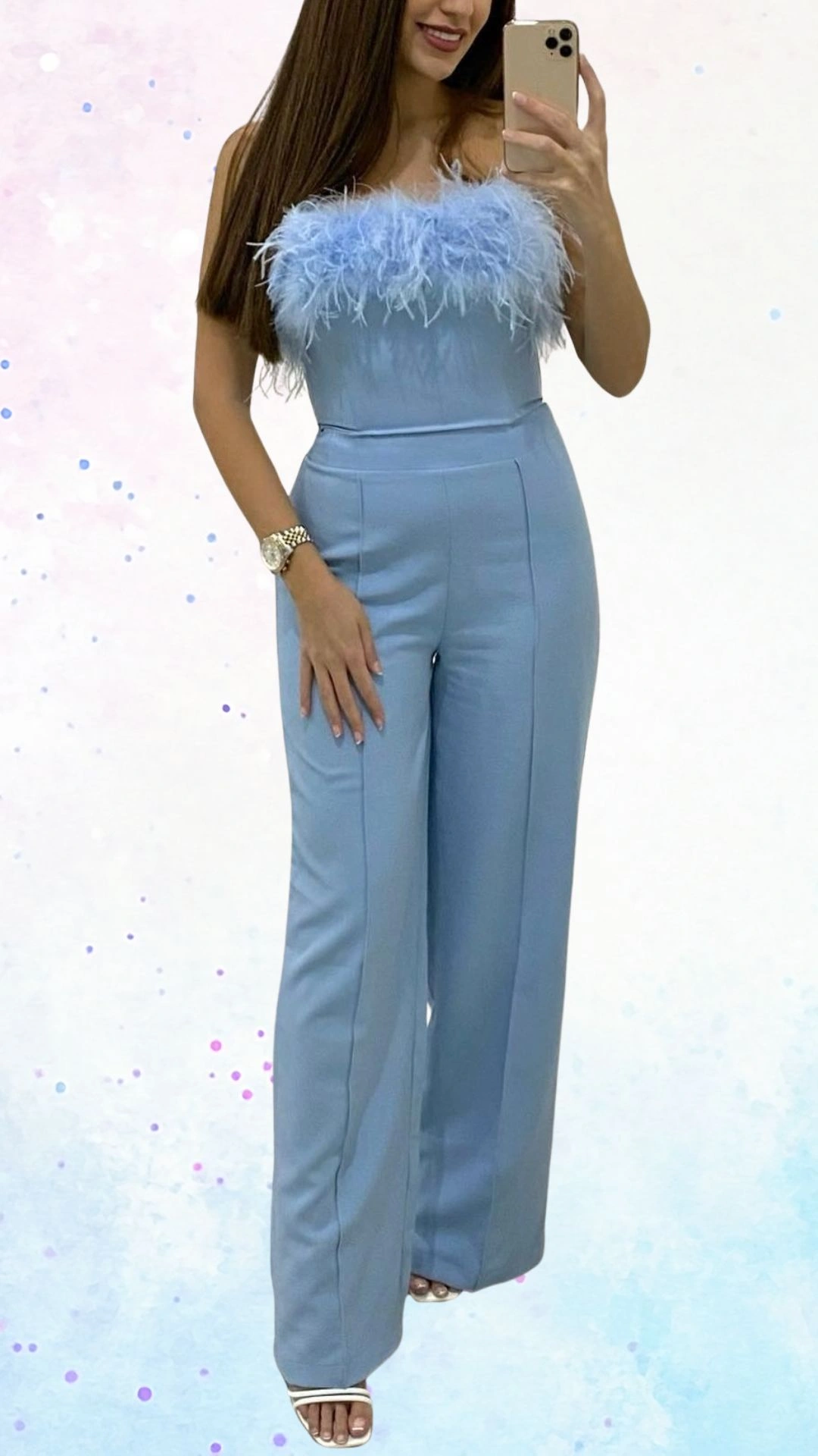 The outfit features a light blue jumpsuit with a strapless design, adorned with a soft, feathery trim at the neckline, adding a playful and celebratory touch. It is made from a smooth fabric that creates a sleek silhouette with wide-leg trousers, offering a comfortable fit. The overall design is elegant yet festive, perfect for a baby shower celebration.