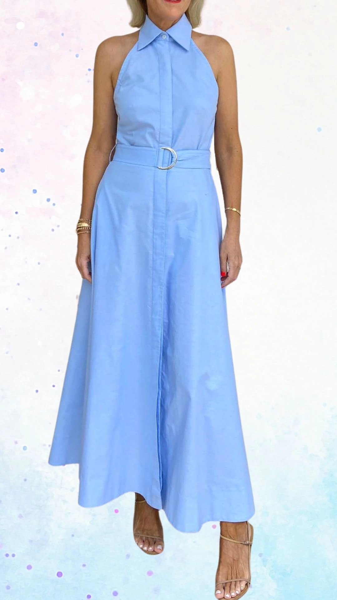 The outfit features a light blue, sleeveless maxi dress made from a soft, smooth fabric that drapes elegantly. It has a button-up front with a collared neckline and a stylish belt with a silver buckle at the waist, adding shape and sophistication to the look. The skirt flows gently, creating a graceful silhouette ideal for a baby shower celebration.