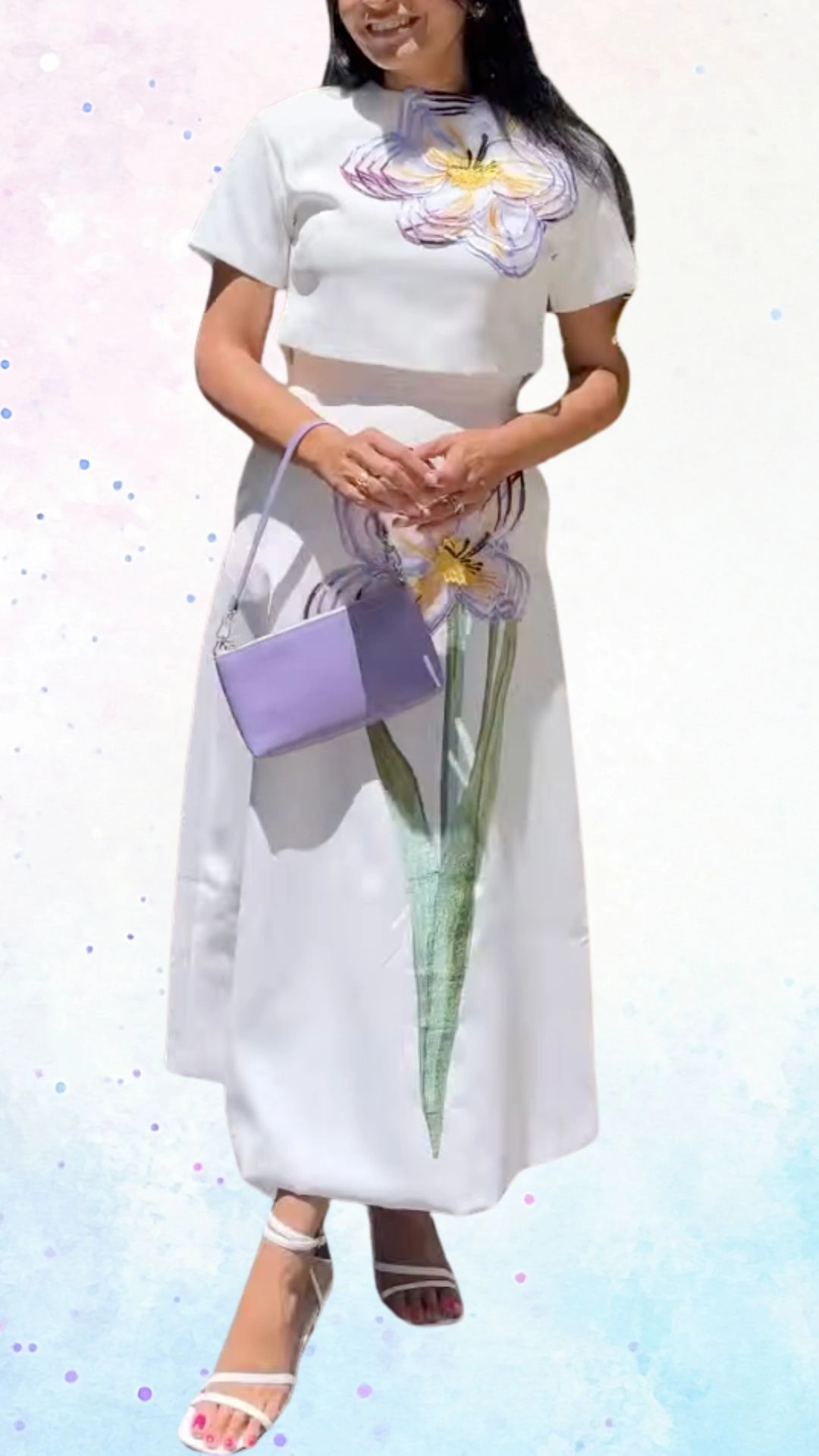 The outfit features a white, fitted crop top with short sleeves, adorned with colorful flower embroidery, transitioning into a floor-length skirt that showcases a vibrant floral design. The lightweight fabric drapes elegantly, complimented by a sleek, pastel purple handbag and matching strappy sandals, making it a cheerful choice for a baby shower celebration.