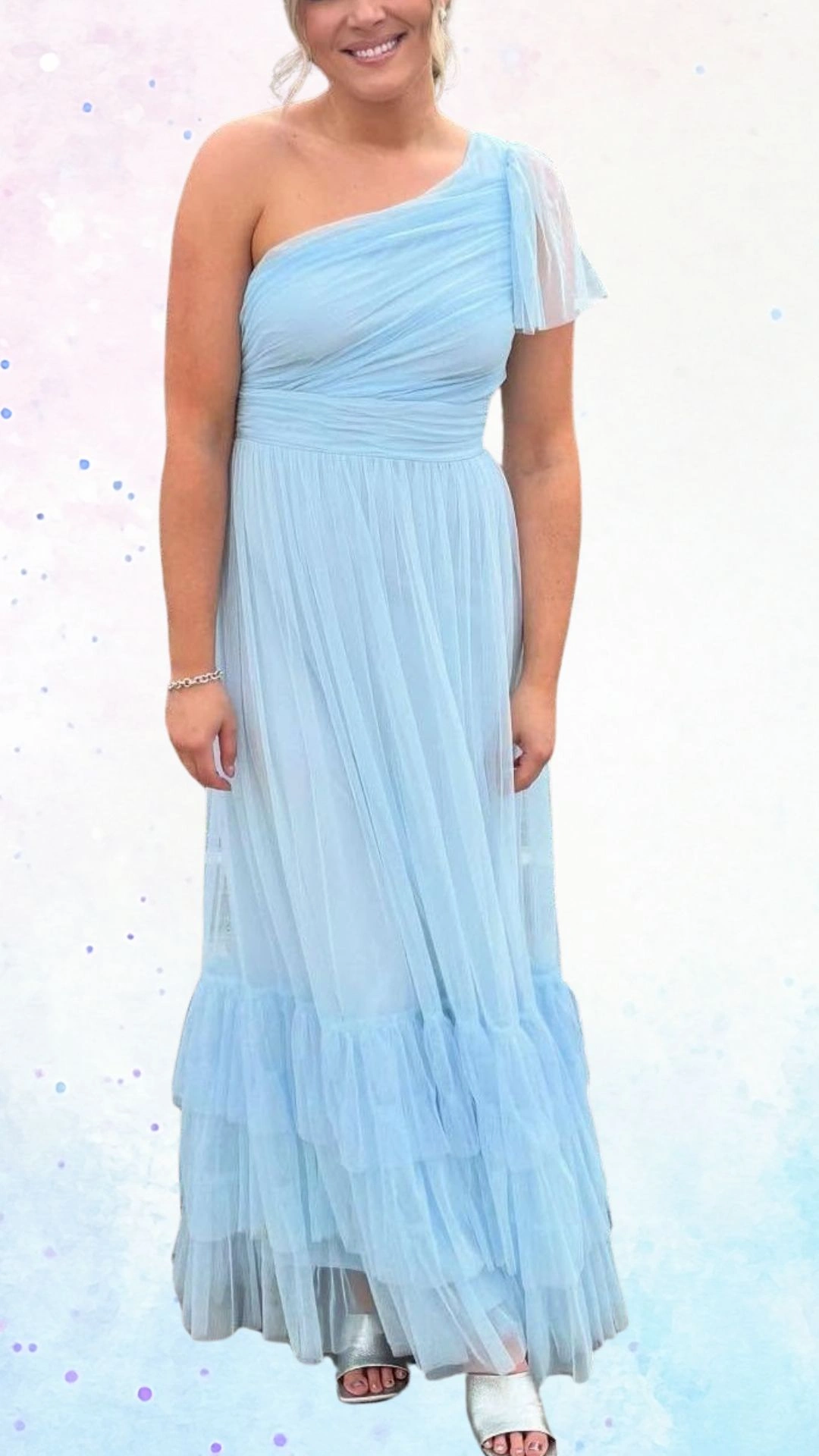 A flowing, one-shoulder dress in soft pastel blue features delicate pleats and a layered ruffle hem, creating an elegant and festive look. The lightweight fabric drapes gracefully, complemented by a sheer, textured sleeve that adds a playful touch, making it a charming choice for a baby shower celebration.