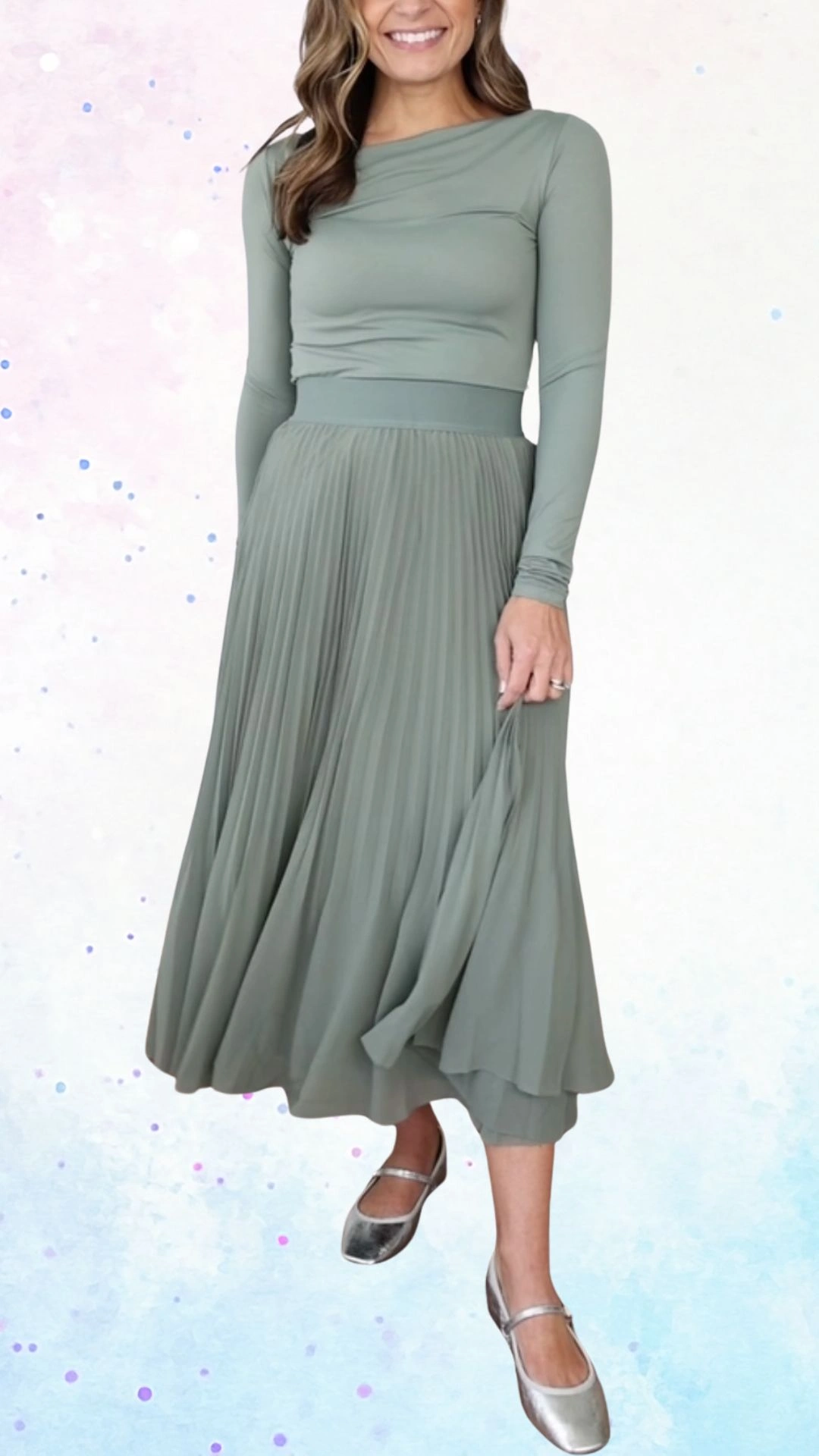 The outfit features a long-sleeved, soft sage green top with a draped neckline, paired with a flowing, pleated midi skirt that adds movement and elegance. The lightweight fabric creates a gentle shimmer, complemented by silver flats that enhance the overall graceful look, making it perfect for a baby shower celebration.