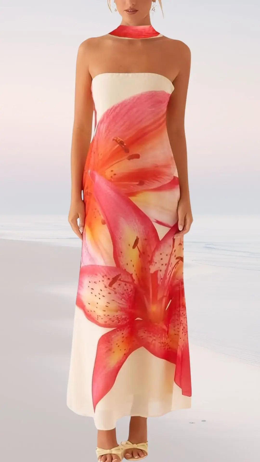 A strapless, floor-length dress features vibrant red and orange lily floral patterns cascading down the fabric, complemented by a matching choker. The dress has a soft, flowy design perfect for a summer beach wedding. Worn with light-colored sandals, it embodies elegance and a tropical aesthetic.