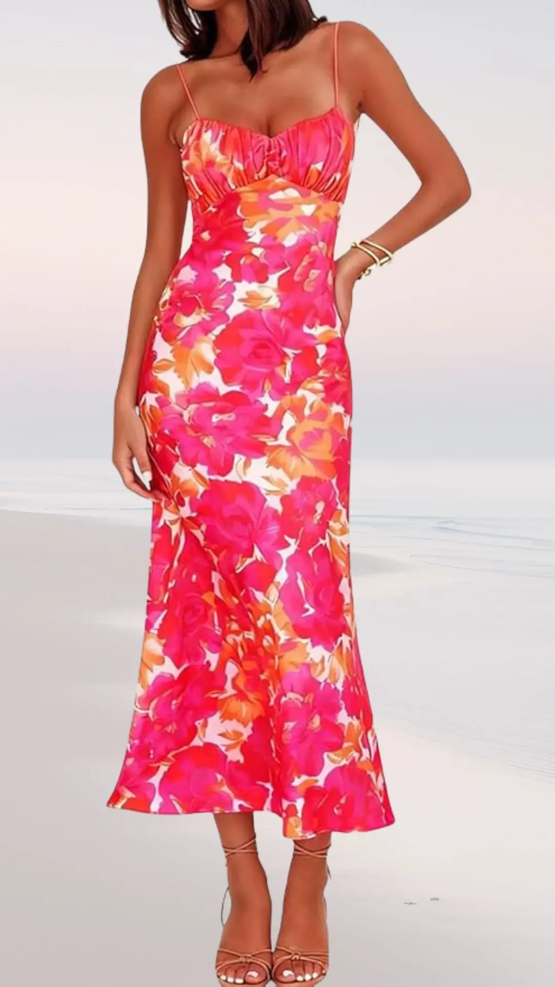 A figure-hugging maxi dress features a vibrant floral pattern in shades of pink and orange. It has thin spaghetti straps and a gathered bodice, gracefully flowing down to a flared hem. The model pairs the dress with nude strappy sandals, set against a soft beach backdrop.