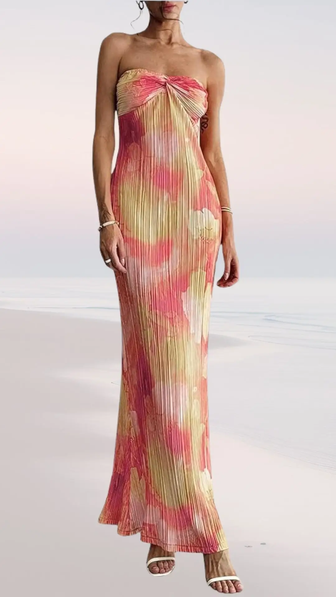 A strapless, floor-length dress features a flowing pleated design in soft shades of pink, yellow, and cream, evoking a beachy, summer vibe. The gown has a fitted bodice that gracefully transitions into a relaxed skirt, perfect for a wedding setting. Complemented by simple white sandals, it embodies elegance and comfort.