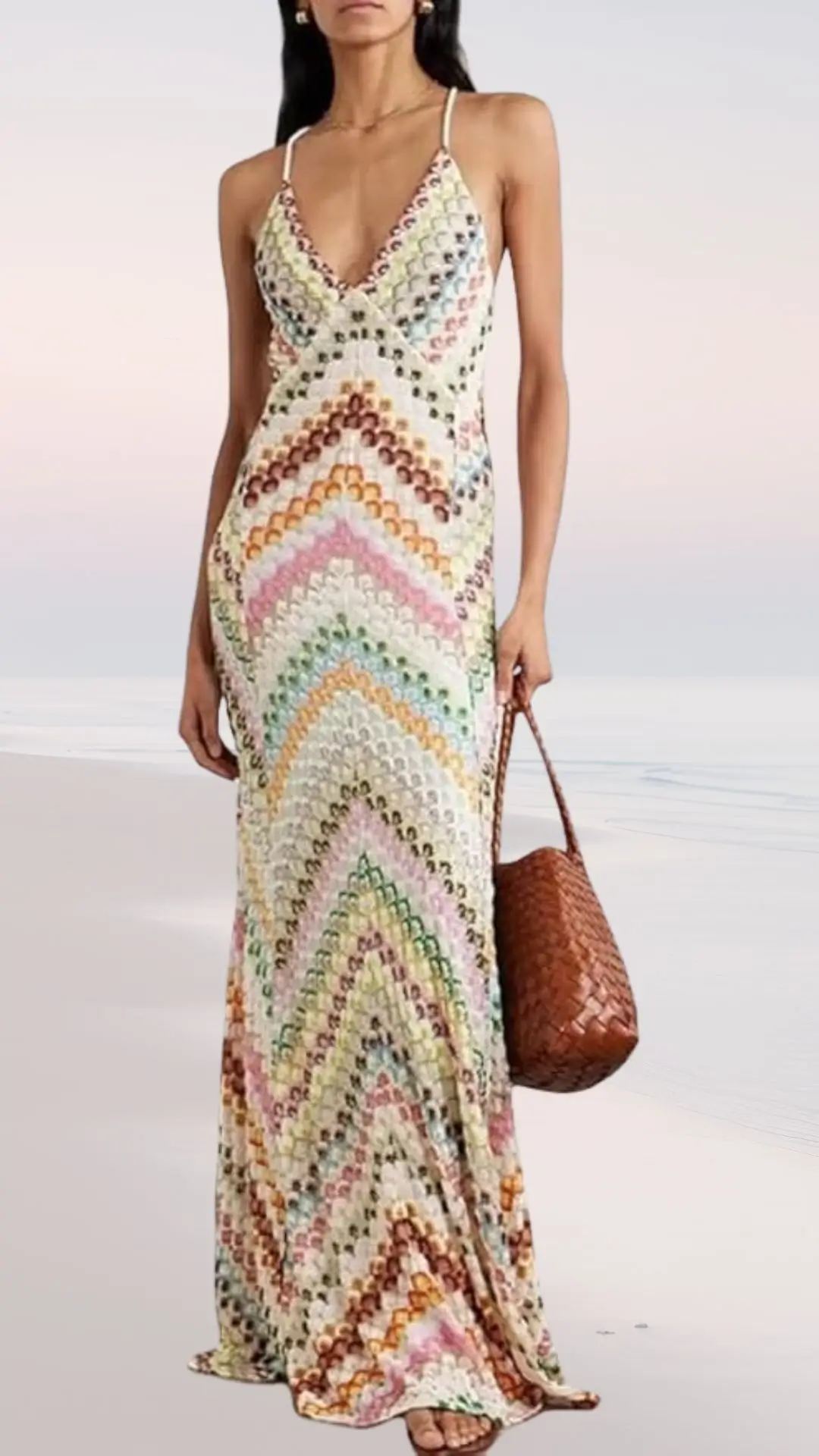 A woman stands on a beach wearing a long, flowy dress featuring a colorful zigzag pattern in pastel shades of pink, green, and brown. The dress has a deep V-neck and thin straps, complemented by a woven brown handbag and strappy sandals. The background hints at a serene beach setting with soft, muted colors.