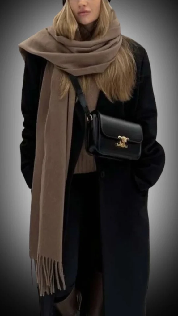 The outfit features a long black overcoat with a sleek, tailored silhouette, layered over a fitted dark top. A soft, beige scarf is wrapped around the neck, adding warmth and texture, while a small black crossbody bag with gold hardware enhances the chic appeal. Paired with dark trousers and subtle brown ankle boots, this ensemble offers an elegant, cohesive look for winter.