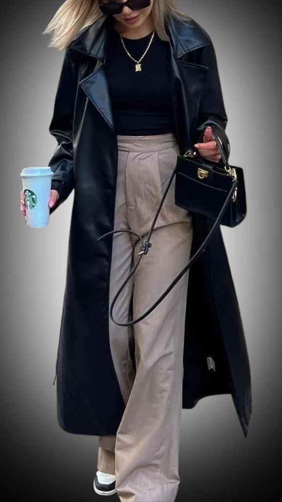 The outfit features a long, black leather duster coat with a sleek finish, layered over a fitted black top. Complementing the coat, the model wears light beige wide-leg trousers that add a stylish contrast, paired with casual black and white sneakers for a relaxed look. Accessories include a small black bag and sunglasses, enhancing the overall chic aesthetic.