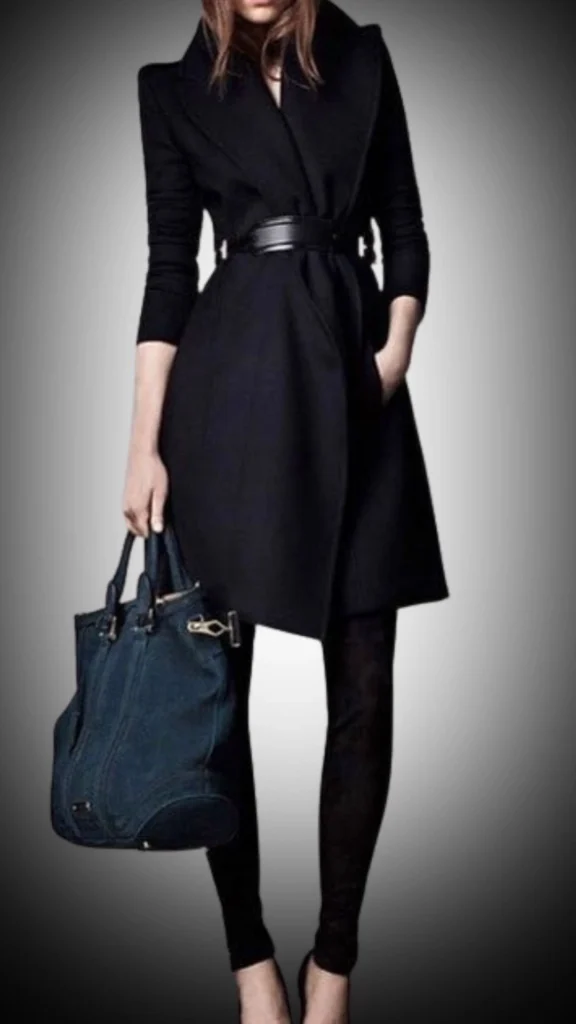A stylish, fitted black coat features a wide, dramatic collar and a defined waist accentuated by a sleek black belt, falling to mid-thigh length. Paired with form-fitting black leggings and elegant black heels, this outfit is complemented by a textured dark blue handbag, creating a chic contrast. The combination of smooth and structured fabrics adds an upscale touch, perfect for winter wear.