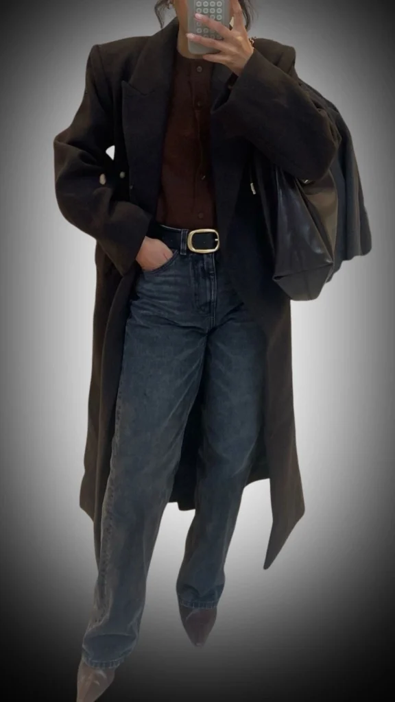 The outfit features a long, stylish black coat with a wide lapel and a relaxed fit, layered over a deep brown button-up top for a rich color contrast. The ensemble is completed with high-waisted, dark denim jeans and pointed-toe heels, creating a polished, contemporary look suitable for winter. Textures in the coat provide warmth while the clean lines and minimalist accessories enhance overall sophistication.