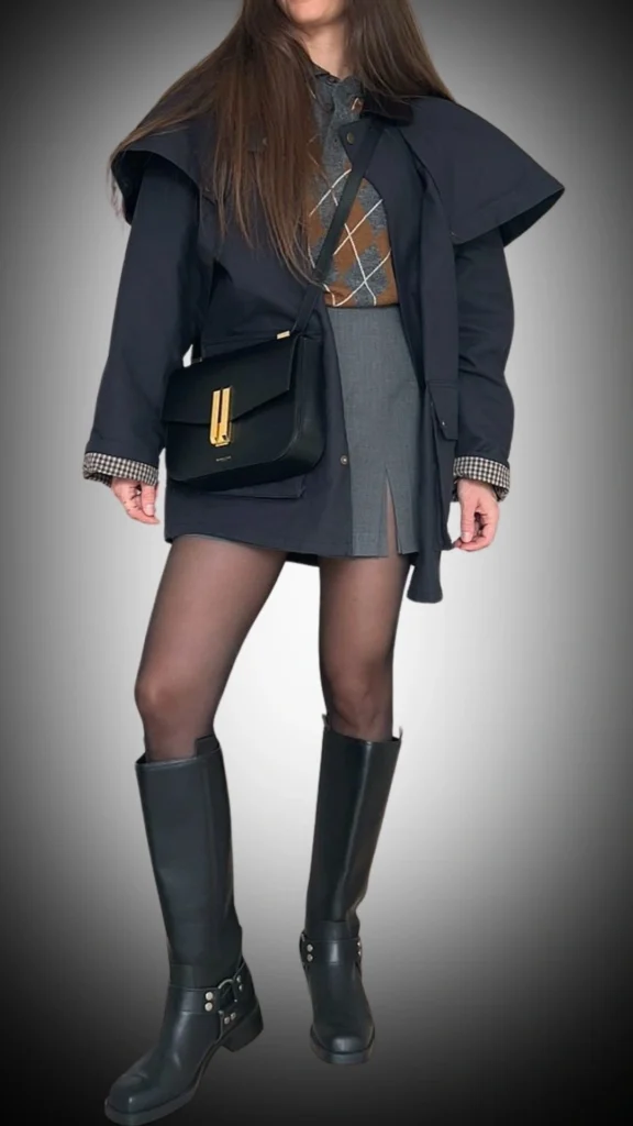 A stylish black coat with a structured silhouette features wide lapels and a relaxed fit, layered over a gray and brown argyle sweater and a gray mini skirt with a subtle slit. The ensemble is completed with sleek black knee-high boots and a chic black crossbody bag with golden accents, creating a polished winter outfit that's both fashionable and functional. The combination of textures and colors adds depth to the look, making it suitable for various occasions.