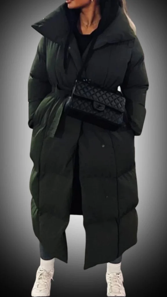 The outfit features a long, oversized dark green puffer coat with a high collar and a belted waist, creating a stylish silhouette. Underneath, a fitted black top complements the coat, while slim dark jeans and classic white sneakers complete the look. The combination of textures and colors adds a modern touch, perfect for winter styling.