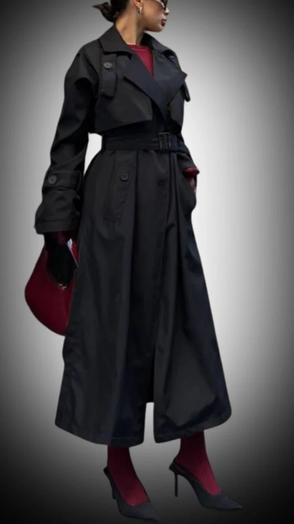 A stylish black trench coat with a belted waist and oversized lapels features a sleek, water-resistant texture that falls below the knees. Underneath, a deep red top complements the coat, paired with vibrant red ribbed socks and pointed-toe black heels, creating a chic and polished winter look. The ensemble is accentuated by a burgundy handbag, adding a pop of color to the monochromatic outfit.