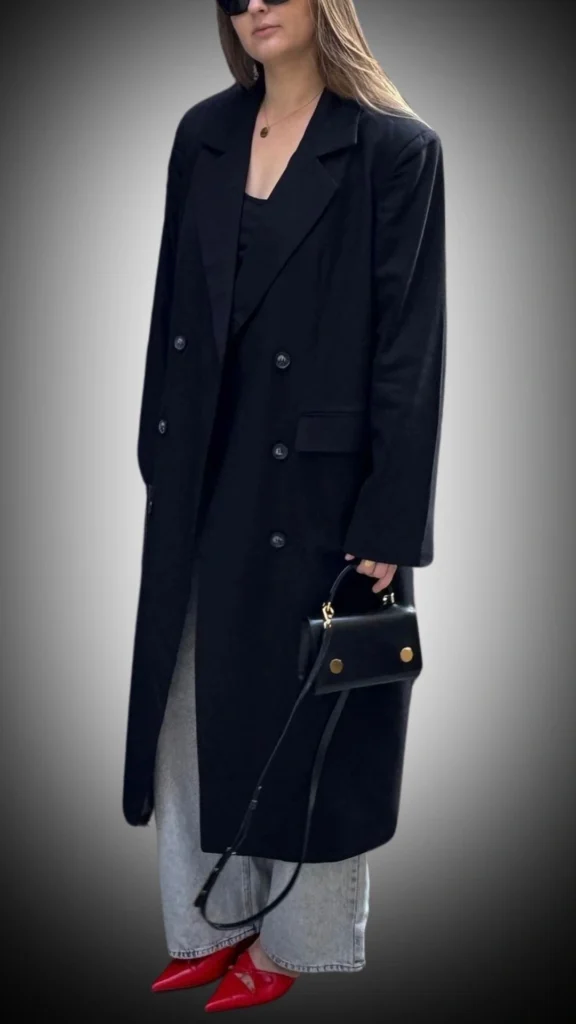 The outfit features a long black double-breasted coat made from a soft, luxurious fabric, providing warmth and style for winter. Underneath, a simple black top is paired with light gray wide-leg jeans, creating a balanced silhouette, while vibrant red pointed-toe heels add a bold pop of color. The look is completed with a sleek black handbag adorned with gold accents, enhancing the overall chic aesthetic.