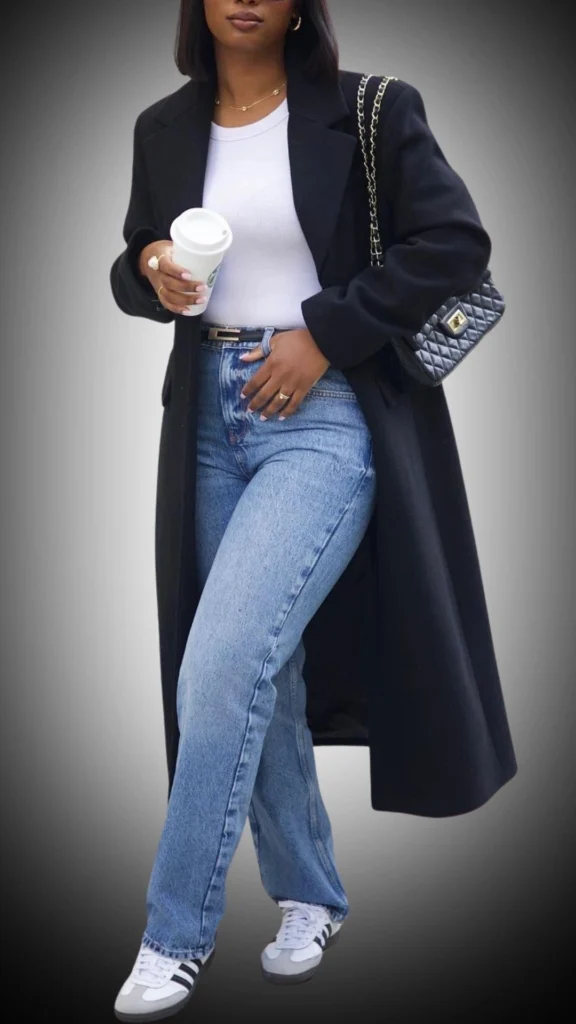 The outfit features a long, black wool coat with a structured silhouette, layered over a fitted white ribbed long-sleeve shirt. Paired with high-waisted, light blue jeans and classic white sneakers accented with black stripes, this look exudes a modern and casual style, perfect for winter wear. A chic quilted handbag and delicate gold jewelry complete the ensemble, adding a touch of elegance.