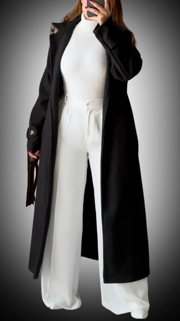 The outfit features a long, tailored black coat with a sleek silhouette, complemented by a ribbed white turtleneck and wide-legged white trousers that create a striking contrast. The coat’s fabric has a smooth texture, while the turtleneck adds warmth, making this ensemble both stylish and functional for winter. Subtle details, like the coat’s buttoned cuffs, enhance its overall sophisticated appearance.