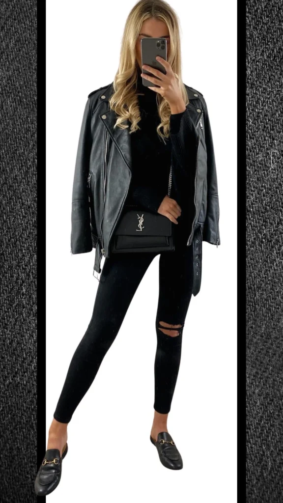 The outfit features form-fitting black jeans with distressed detailing, paired with a chunky black knit sweater for warmth. A sleek black leather biker jacket adds a touch of edge, complemented by stylish black loafers adorned with gold hardware and a matching black handbag for a polished finish.