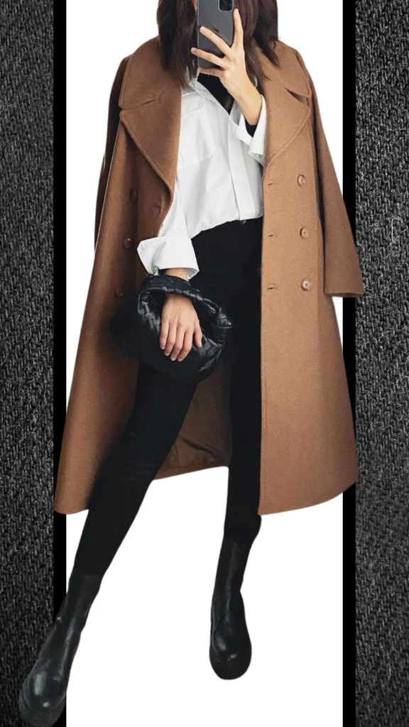 A stylish winter outfit features fitted black jeans paired with a light, oversized white button-up shirt, and a warm, caramel-colored coat draped loosely over the shoulders. The look is complemented by sleek black ankle boots and a compact black handbag, creating a chic contrast in textures and colors while ensuring warmth and comfort for cooler months.