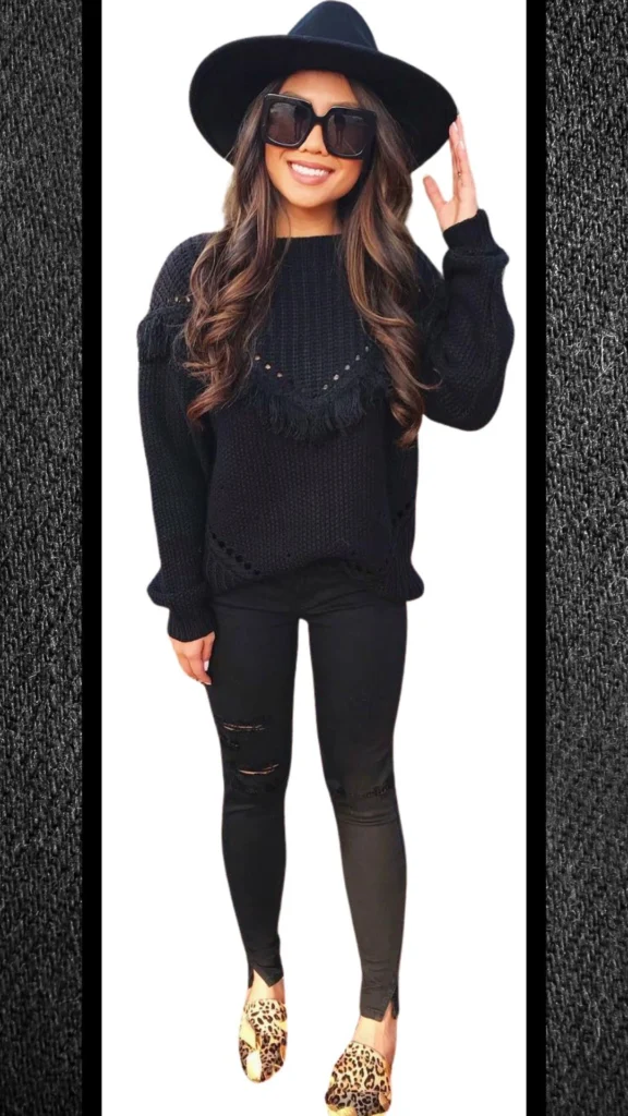The outfit features a cozy black knit sweater with a textured finish, adorned with fringe detailing and open knit accents. Paired with form-fitting black jeans that have subtle distressing, the look is completed with stylish leopard print slip-on shoes and oversized black sunglasses. A wide-brimmed black hat adds an elegant touch, making it a chic and comfortable ensemble for colder months.