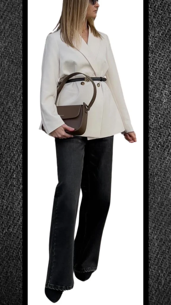 The outfit features high-waisted, black jeans with a slightly flared cut, creating a sleek silhouette. Paired with a structured, cream-colored blazer that has a cinched waist accentuated by a black belt, the ensemble balances warmth and sophistication. Accessories include stylish sunglasses and a minimalist brown crossbody bag, adding a touch of elegance to the winter look.