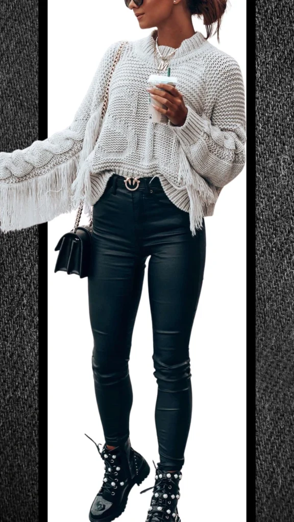 The outfit features a textured, light gray knit sweater with fringe details, paired with form-fitting black jeans that have a sleek, shiny finish. Accessories include a small black handbag and chunky black lace-up boots adorned with silver accents, creating a stylish contrast perfect for cooler months. The overall look combines warmth with a touch of modern edge, ideal for winter outings.