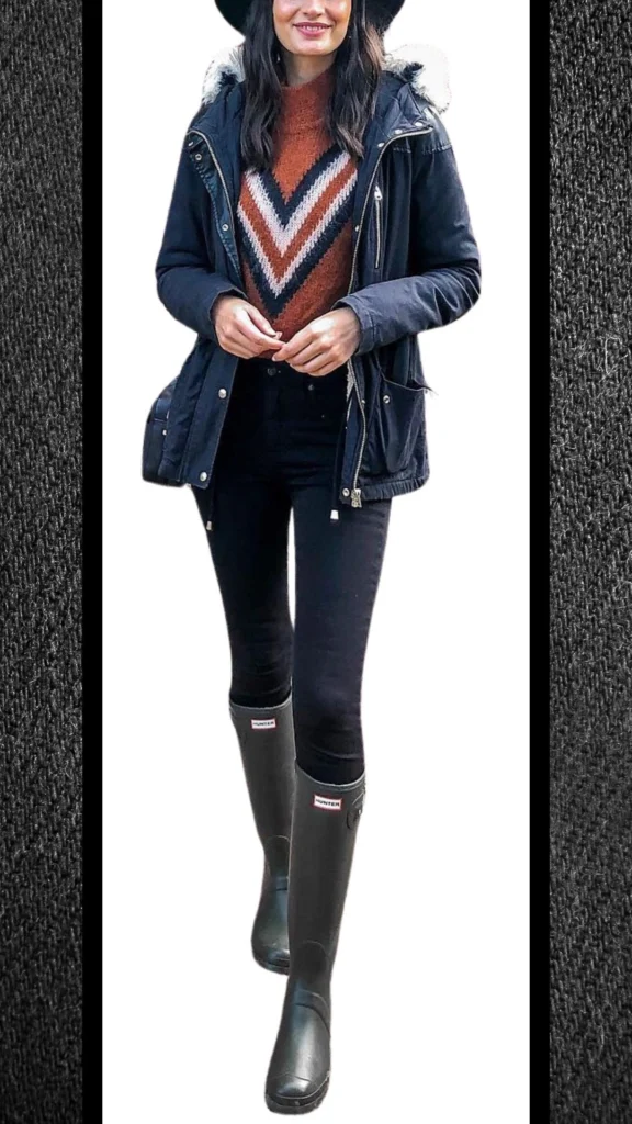 A warm, navy parka jacket features a faux fur-lined hood, paired with a cozy rust and cream chevron-patterned sweater that adds texture. The outfit includes sleek black high-waisted jeans, complemented by stylish black rain boots with subtle branding, creating a chic and functional winter look.