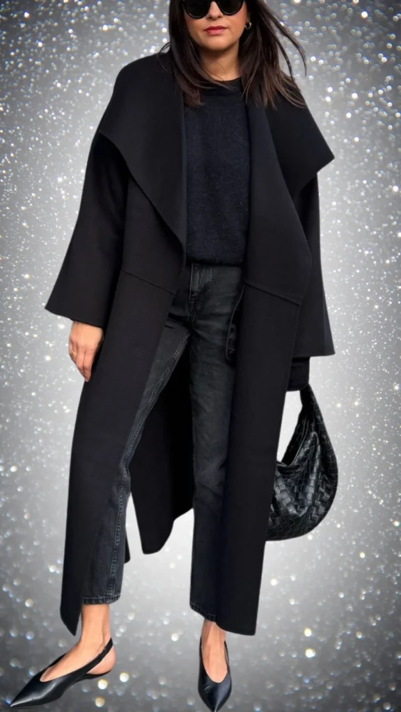 A chic all-black winter outfit features a long, flowing black coat crafted from a soft, textured fabric layered over a cozy black knit sweater. The ensemble is complemented by tailored black jeans and sleek black slingback shoes, while a stylish woven black handbag adds a touch of sophistication. Bold black sunglasses complete the look, enhancing its modern elegance.