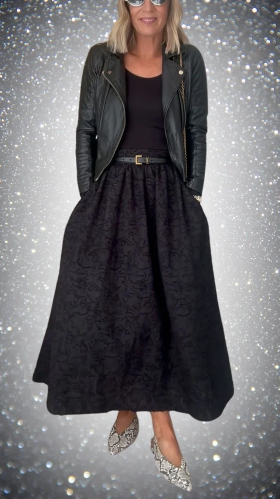 The outfit features a fitted black leather biker jacket layered over a sleek black top, paired with a flowing black skirt adorned with a subtle floral texture for added depth. Accessories include a thin black belt cinching the waist and stylish snake-print flats that introduce a contrasting pattern while keeping the monochromatic theme. This ensemble is perfect for winter, combining warmth, elegance, and chic style.