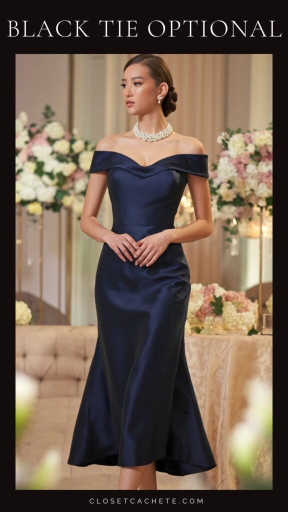 A fitted navy gown features an elegant off-the-shoulder neckline and a flattering A-line silhouette that gracefully flares out at the hem. Complemented by a statement pearl necklace, the outfit epitomizes sophistication suitable for a black tie optional event. The backdrop includes soft florals, enhancing the formal ambiance.