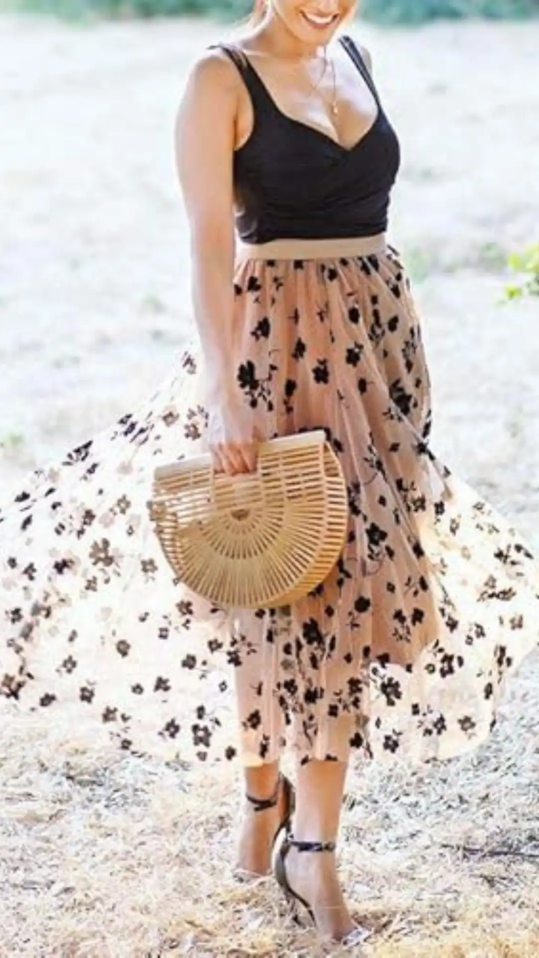 A flowing, high-waisted midi skirt in a soft, sheer fabric features a delicate blush base adorned with bold black floral patterns and is paired with a fitted black crop top, creating a chic silhouette. The look is completed with a stylish wooden handbag and strappy high-heeled sandals, making it perfect for a romantic outdoor event or casual gathering.