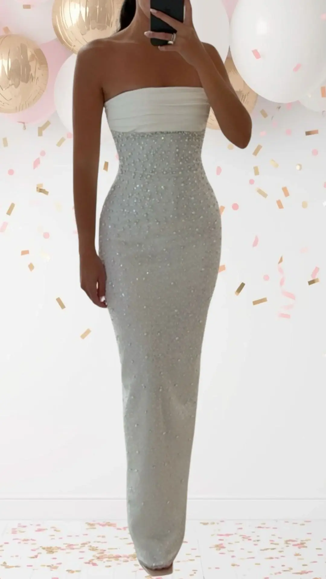 The bridal shower outfit features a fitted, strapless gown with a delicate off-white bodice and a shimmering, silver sequined skirt. The sleek fabric cascades to the floor, adorned with subtle beadwork that adds elegance and a touch of whimsy without being overly formal. The dress combines sophistication with celebratory charm, making it ideal for a festive occasion.