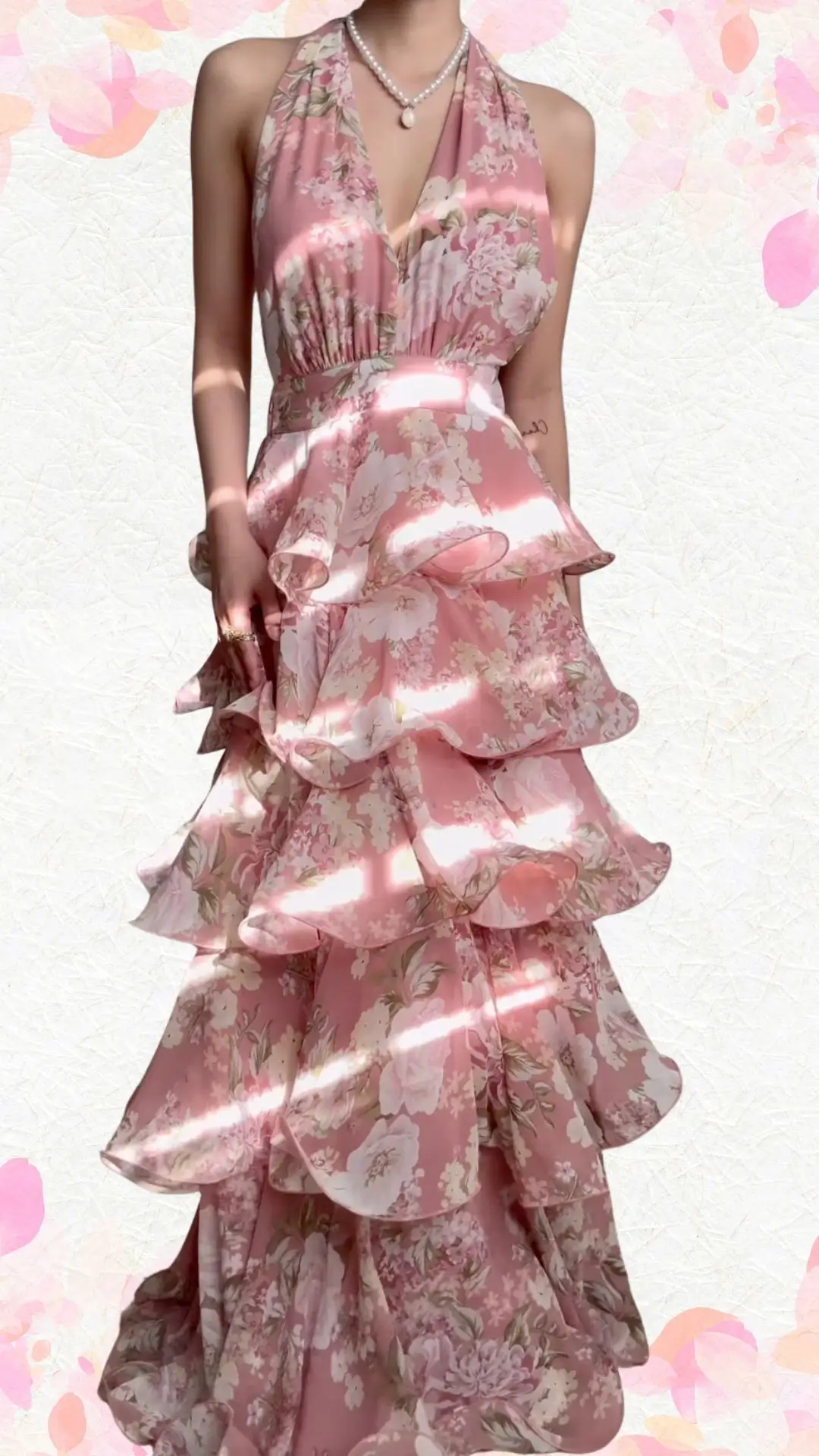 The outfit features a halter-neck design in a soft pink fabric adorned with delicate floral prints, creating a playful yet elegant aesthetic. It showcases a fitted bodice with a flowing, tiered skirt composed of cascading ruffles, adding a whimsical touch to the overall look. The deep V-neckline and elegantly draped fabric enhance the outfit's celebratory vibe without appearing overly formal.