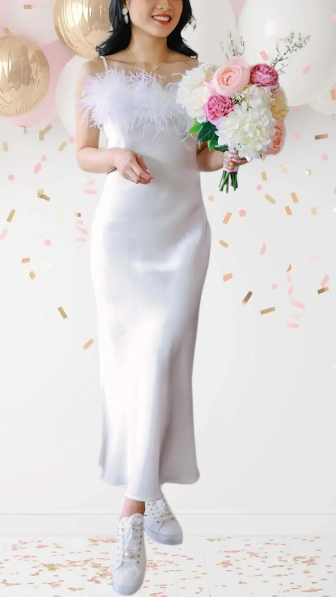 The bride is wearing a sleek, satin midi dress in a soft white shade, featuring delicate feather accents on the shoulders that add a whimsical touch. The dress has a subtle, fitted silhouette with thin straps and a sophisticated scoop neckline, perfectly balanced by casual white sneakers to maintain a celebratory yet playful vibe. This outfit blends elegance with fun, making it ideal for a bridal shower.