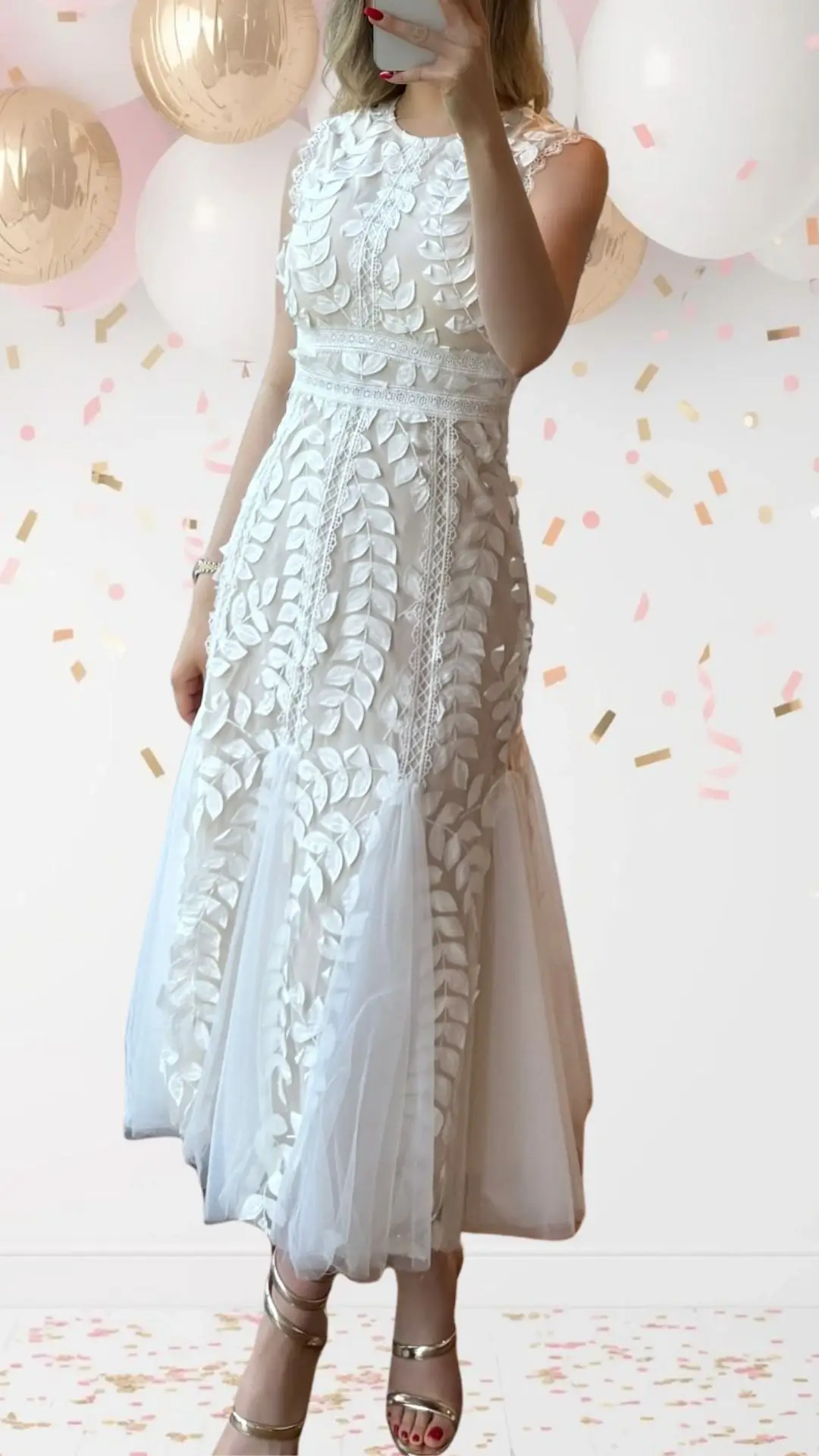 The bridal shower outfit features a sleeveless white dress adorned with intricate three-dimensional leaf appliqués and delicate lace trim along the bodice and waist. The A-line silhouette is complemented by a sheer tulle overlay that flares at the hem, adding a touch of whimsy without compromising elegance. Paired with strappy metallic sandals, this look perfectly balances festivity and refinement.