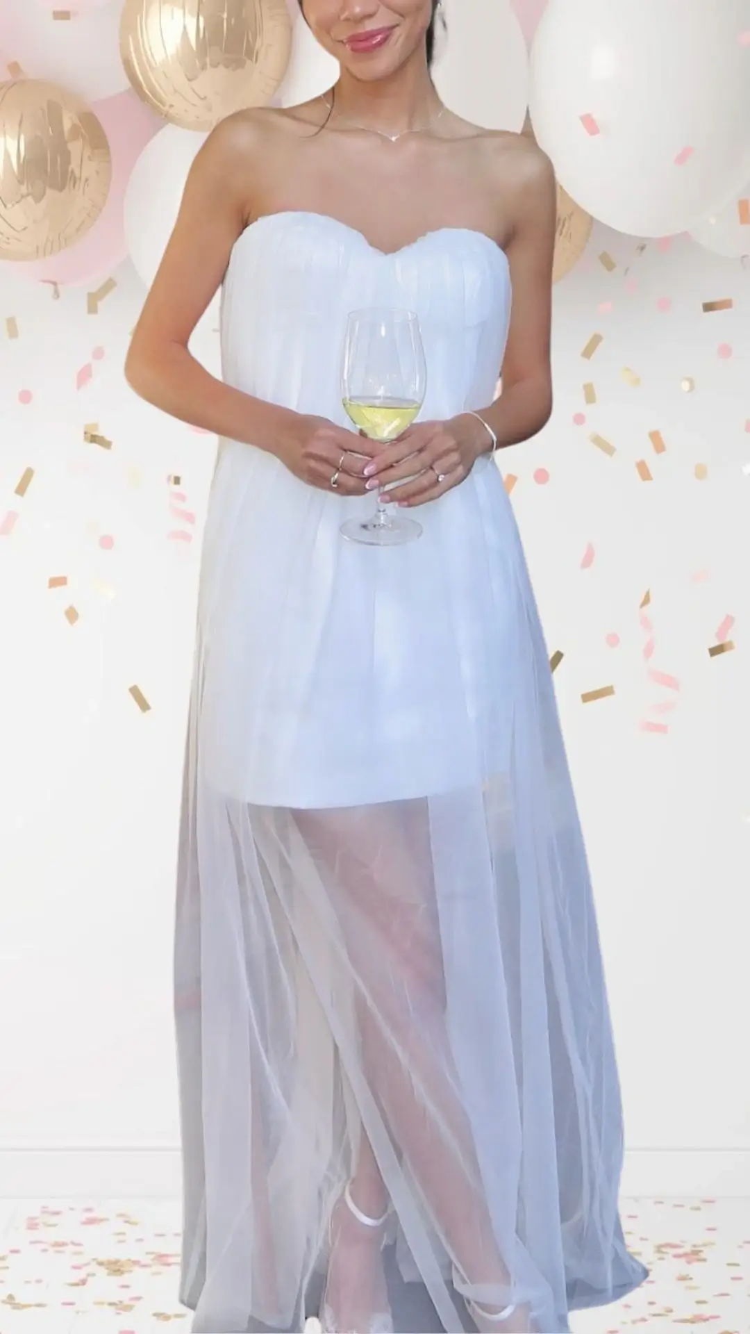 The outfit features a strapless, sweetheart neckline dress in a soft white fabric, complemented by a sheer overlay that adds an ethereal touch. Delicate pleats adorn the bodice, while a flowing skirt with a slightly longer hem creates movement and elegance, offering a playful yet sophisticated vibe. The look is completed with subtle high-heeled shoes that enhance the outfit's overall charm without overwhelming it.