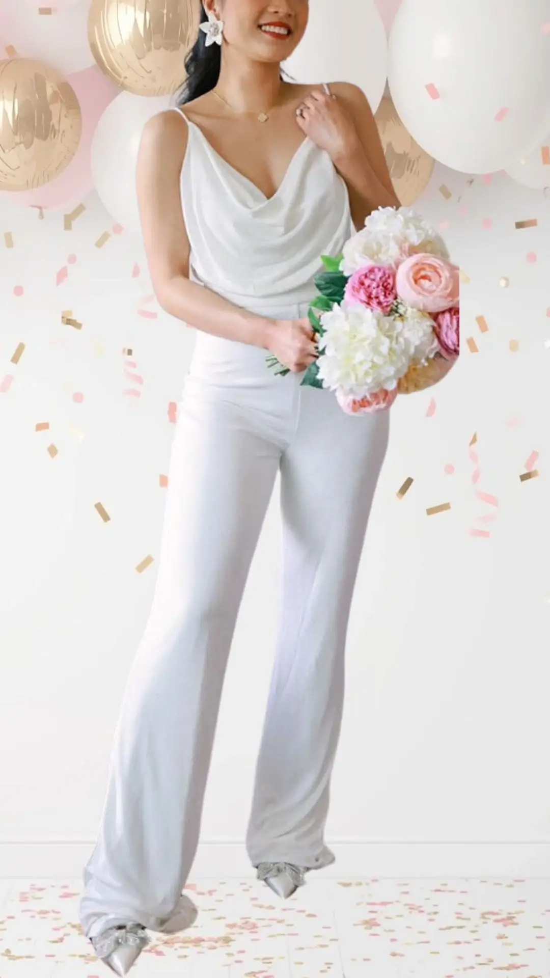The bride wears a sleek, white jumpsuit featuring a draped cowl neckline and slender spaghetti straps. The fabric is smooth and lightweight, elegantly flowing into wide-leg trousers that add a touch of sophistication. Paired with silver, embellished shoes, this outfit blends classic elegance with a playful spirit, perfect for a bridal shower celebration.