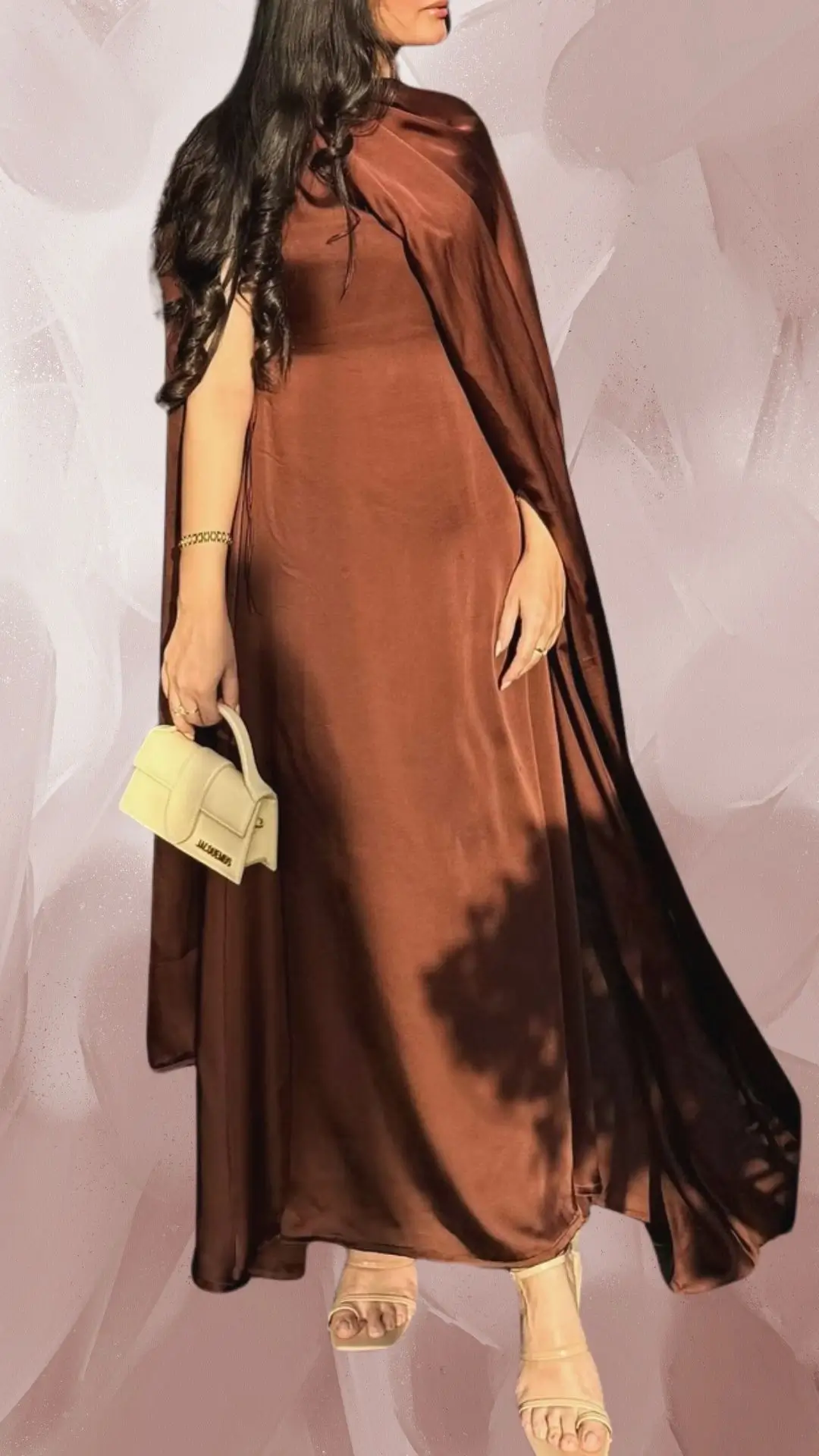 The outfit features a long, flowing brown dress made from a smooth, lightweight fabric with a subtle sheen. It has elegant draped sleeves and a round neckline, paired with neutral-toned strappy sandals that complement the dress's sophisticated look. Completing the ensemble is a small, chic handbag for an added touch of refinement.