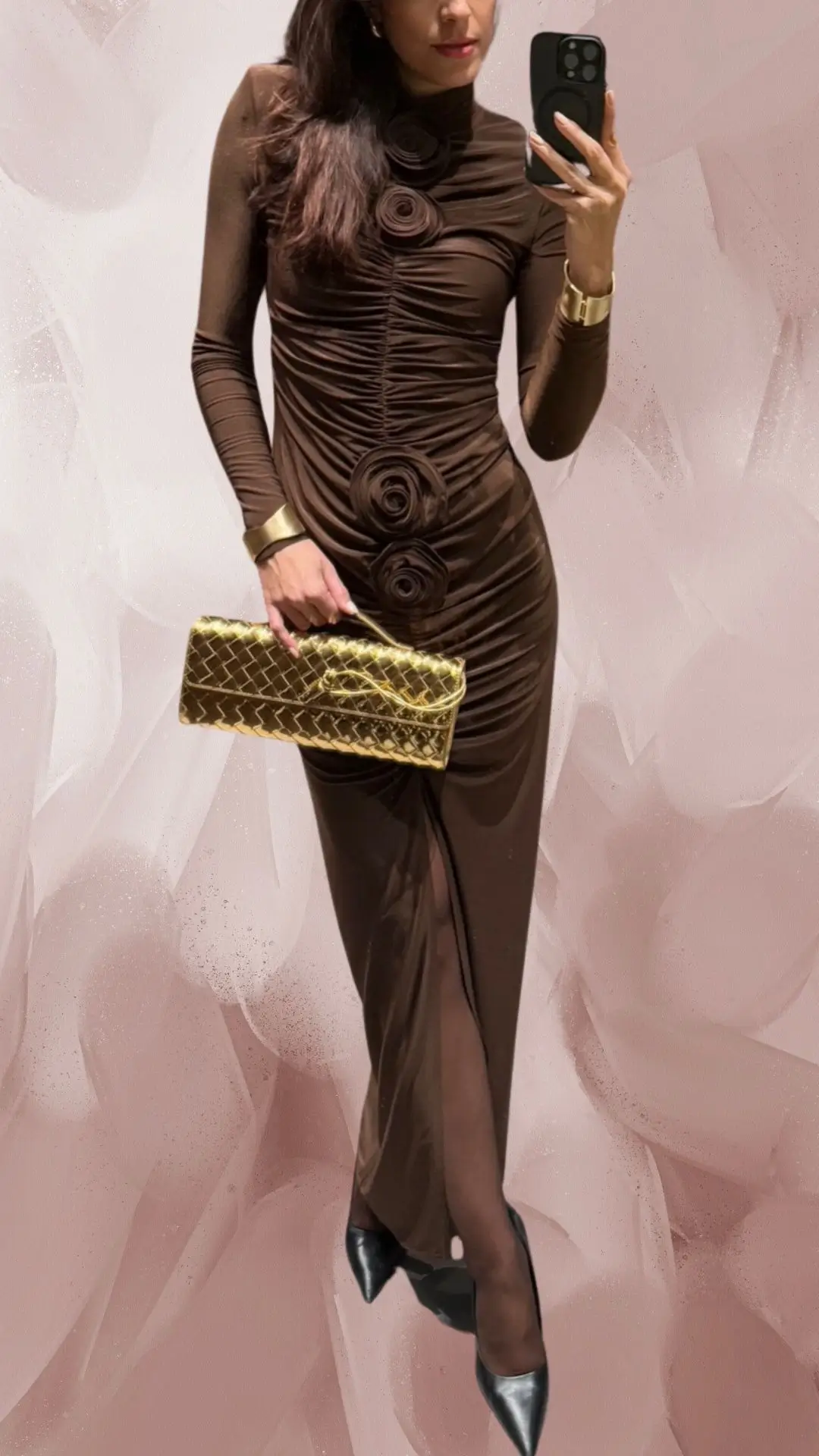 A long, fitted brown dress made from soft, ruching fabric features a high neckline and long sleeves, adorned with floral embellishments down the center. The dress has a subtle side split that enhances its elegant silhouette and is paired with sleek, pointed-toe black heels. Complemented by a gold clutch, this outfit exudes sophistication and style.