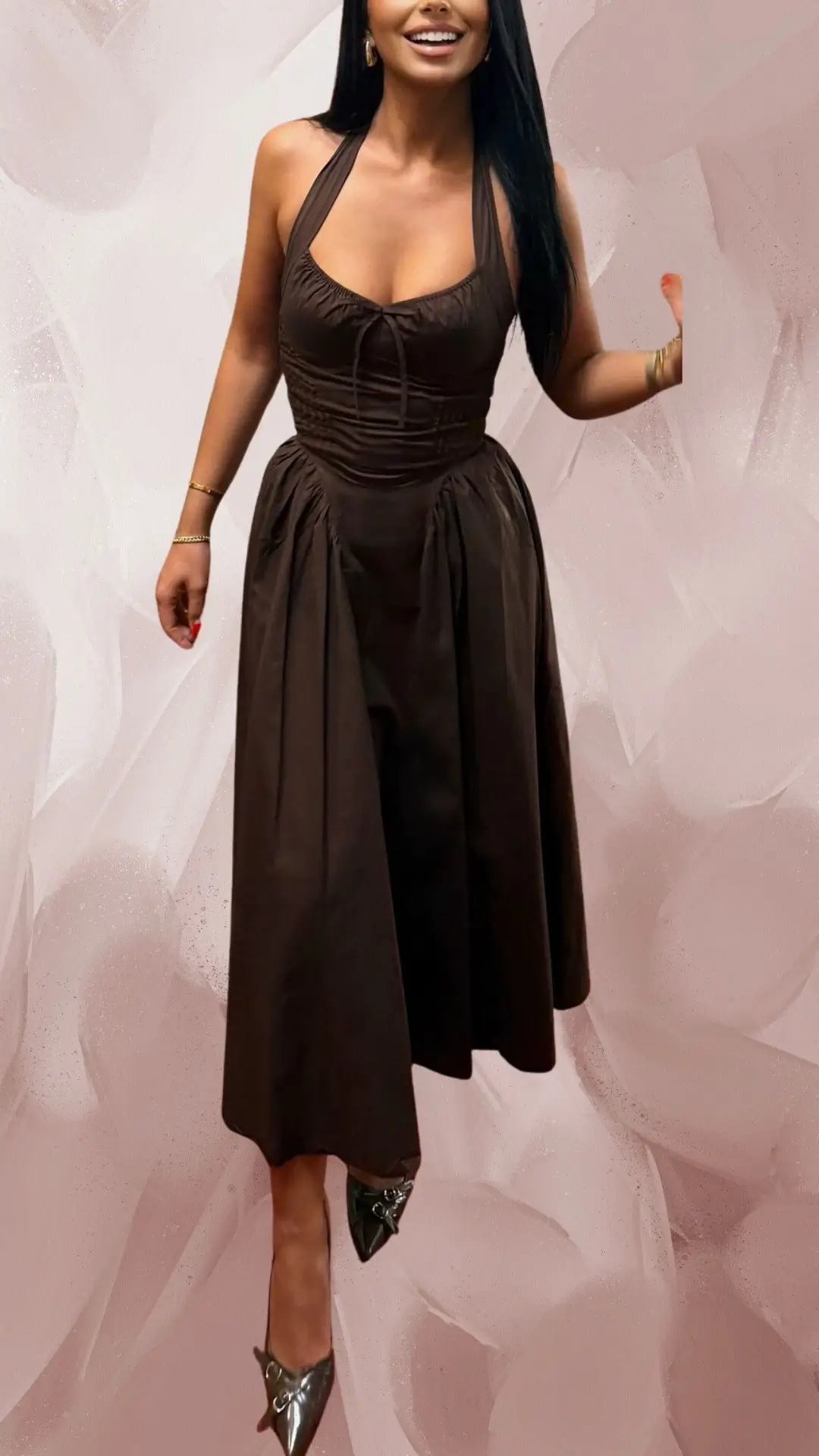 The outfit features a long, brown halter-neck dress made of a soft, flowing fabric with a fitted bodice and a gently gathered, full skirt. It has a sweetheart neckline and is complemented by sleek, pointed-toe shoes in a shiny metallic finish. The overall look exudes elegance, accentuated by the interplay of textures and a sophisticated silhouette.