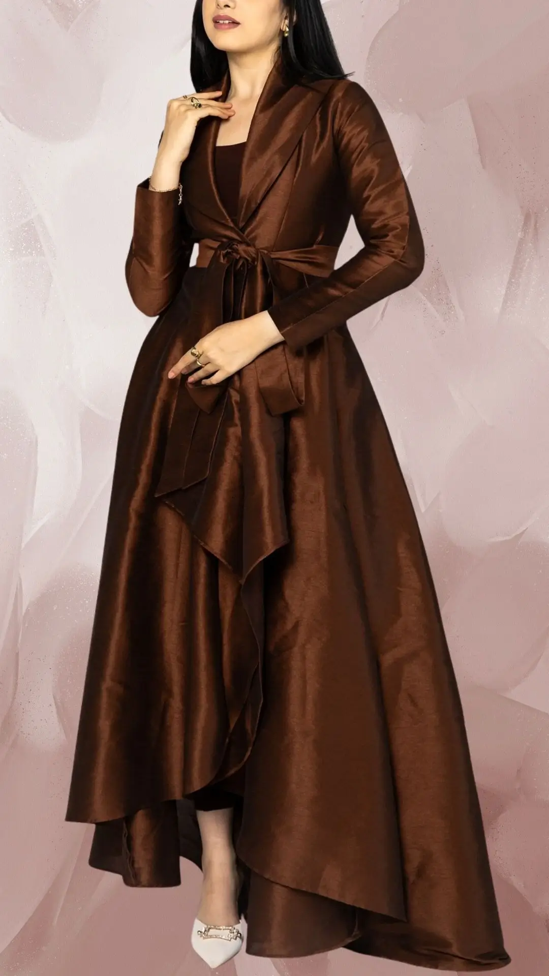 A long, flowing brown dress made from a glossy satin fabric features a fitted bodice with a V-neckline and long, tailored sleeves. The waist is cinched with a matching tie, creating elegant drapes that cascade from the skirt, complemented by chic white pointed-toe heels for a refined finish.