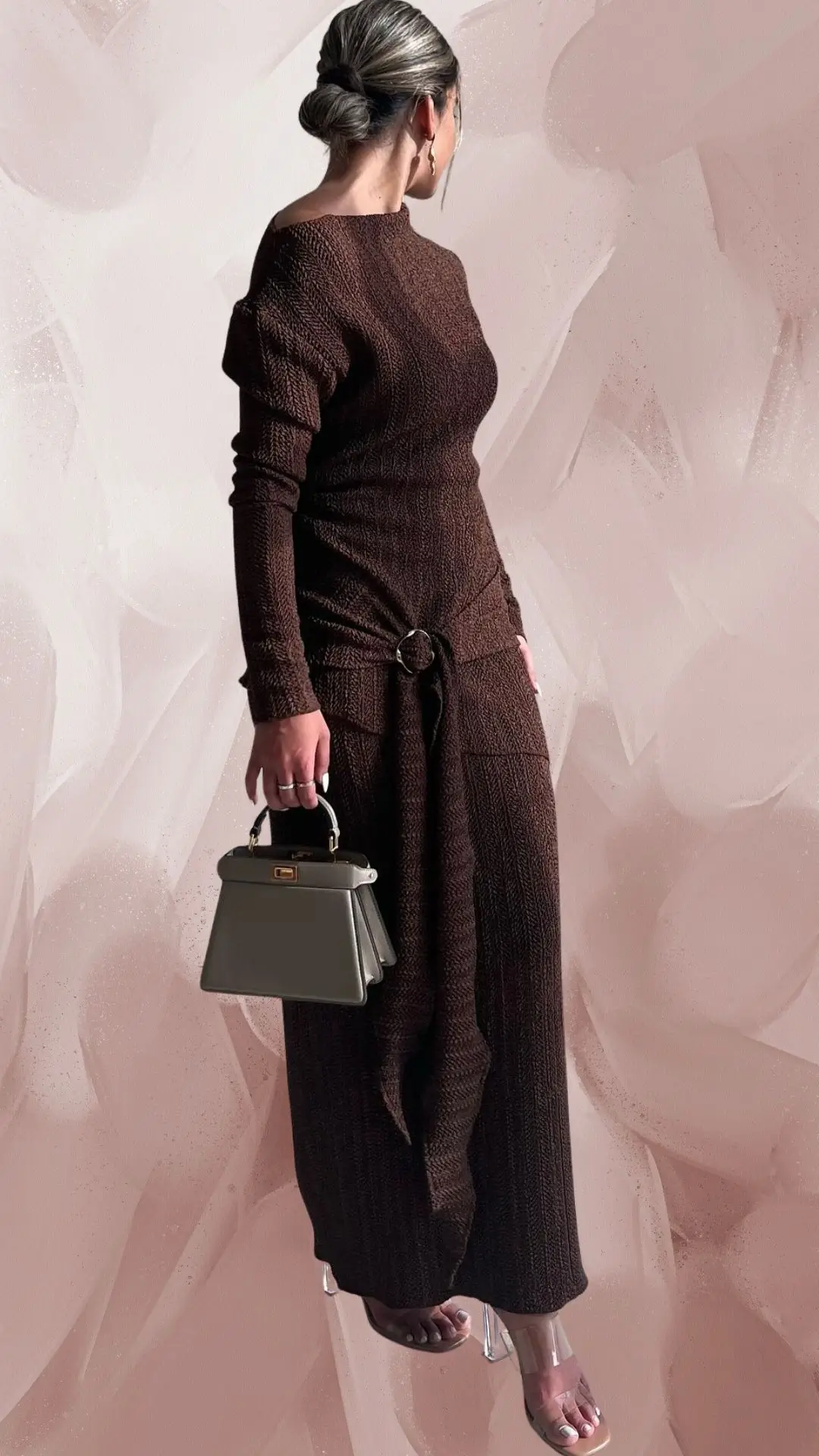 The outfit features a long brown, knit dress with a fitted bodice and a flowing skirt, highlighted by a side knot detail at the waist. It has long sleeves and a scoop neckline, crafted from a textured fabric that adds depth to the rich brown shade. The look is complemented by clear, strappy heels and a sophisticated, gray handbag.