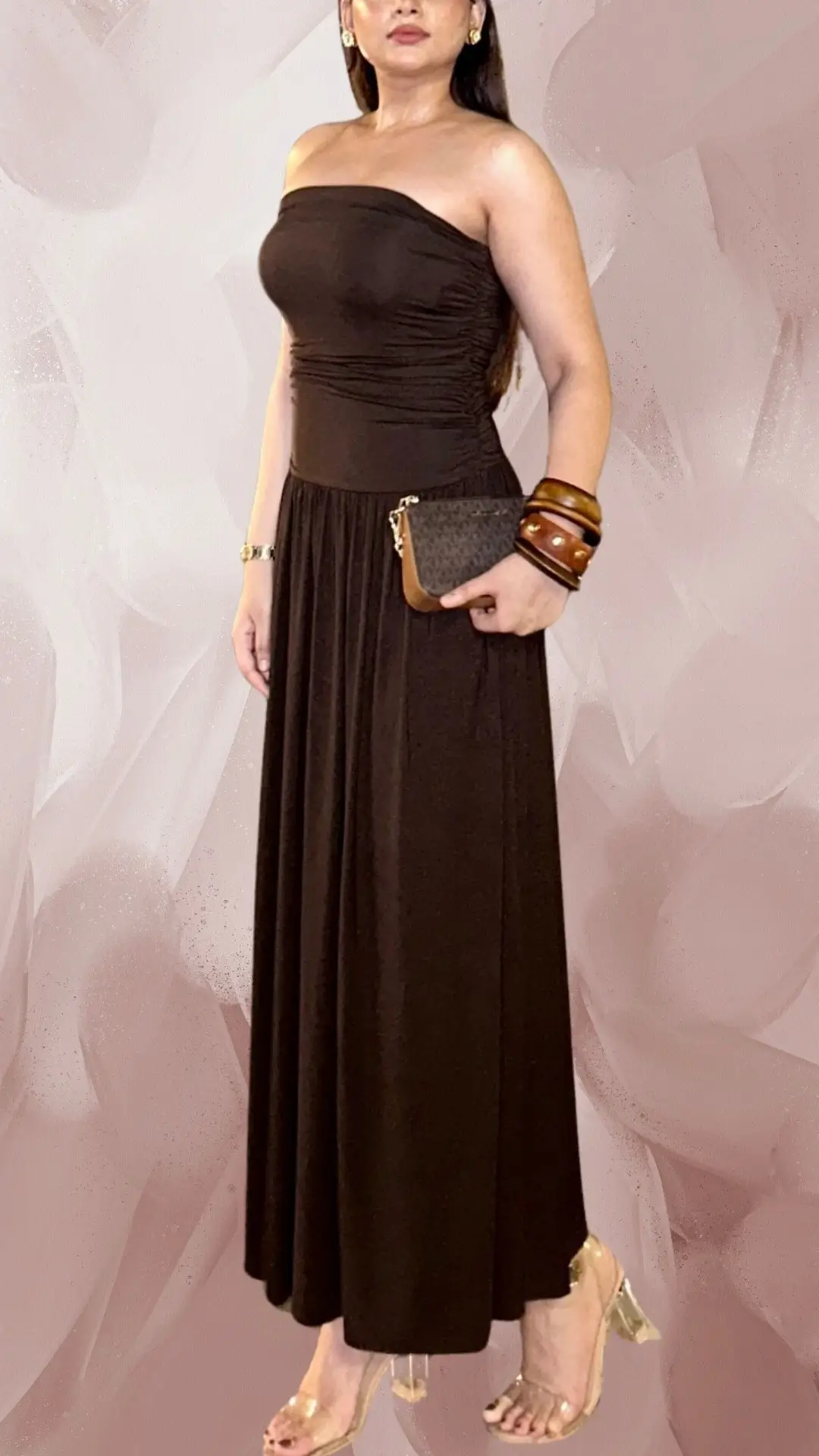 This elegant long brown dress features a fitted, strapless bodice with a flattering silhouette that flows into a gentle A-line skirt. Made from a soft, textured fabric, the gown is complemented by transparent heeled sandals and statement wooden bangles, adding a touch of sophistication to the ensemble. The rich brown shade enhances the overall look, making it suitable for formal occasions.