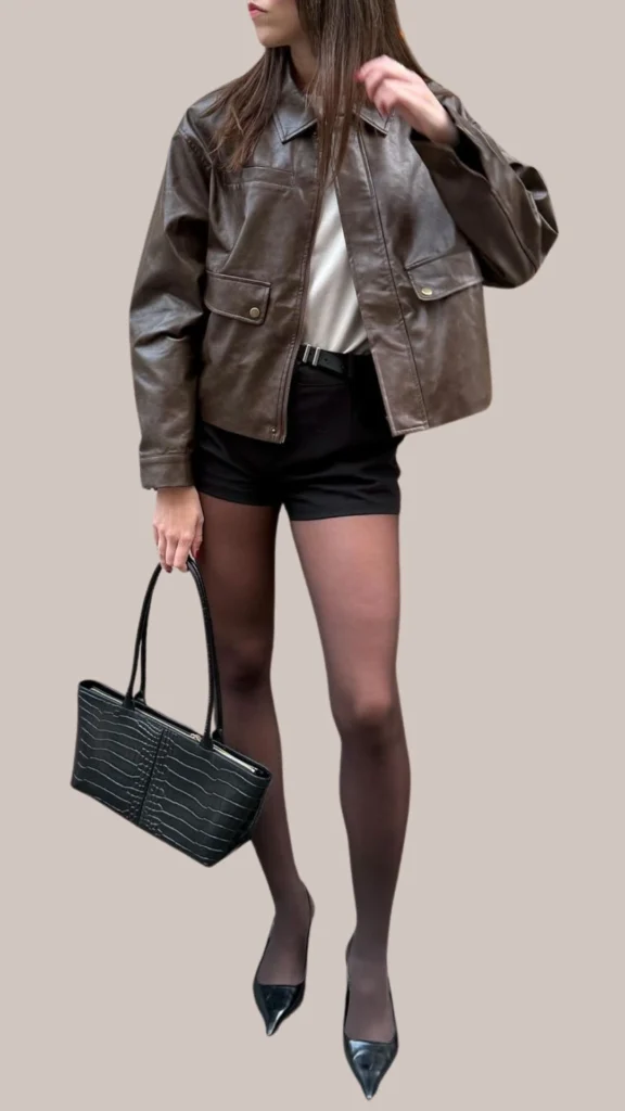 A glossy dark brown leather jacket features a relaxed fit with a collared neckline and two front pockets, layered over a classic white top. This chic outfit includes tailored black shorts and sheer black tights, complemented by sleek black high-heel shoes and a textured black handbag for added sophistication. The combination of rich browns and blacks creates a stylish and versatile look.