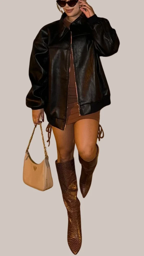 A chic ensemble features a glossy black leather bomber jacket paired with a fitted brown mini dress. The outfit is complemented by knee-high crocodile-embossed brown boots and a small cream-colored handbag with a gold strap, creating a stylish contrast of textures and shades. Dark sunglasses add a fashionable finishing touch.
