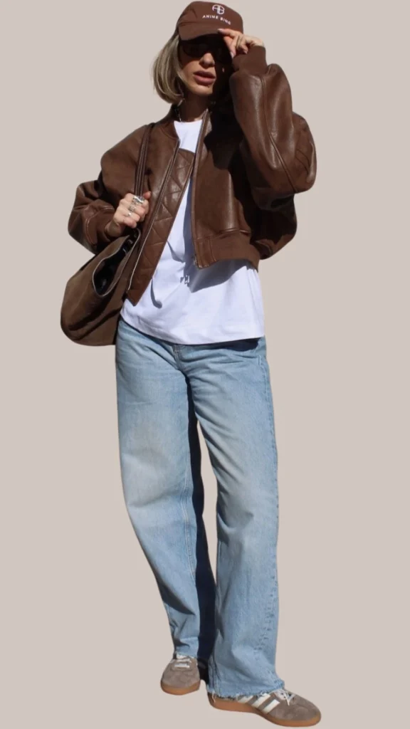 A chic brown leather bomber jacket with a glossy finish is layered over a casual white t-shirt, complemented by light blue, loose-fitting jeans. The outfit is accessorized with a deep brown cap and grey slip-on sneakers featuring white stripes, creating a stylish yet relaxed vibe. The overall texture contrasts the smooth leather with the soft cotton and denim fabrics, enhancing the outfit's cohesiveness.