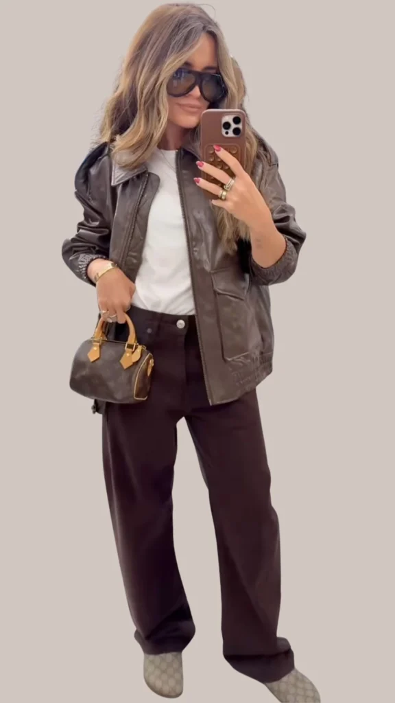 The outfit features a rich, dark brown leather jacket with a sleek, glossy texture, layered over a classic white t-shirt. Paired with wide-leg dark brown trousers, the look is complemented by a small, structured handbag in a matching brown hue. Accessories include chic oversized black sunglasses and subtle jewelry, enhancing the outfit’s sophisticated vibe.
